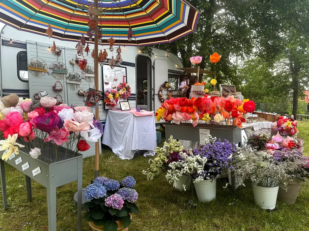 Ein bunt geschmückter Verkaufsstand auf dem Kunsthandwerkermarkt "Fair liebt Kunst" in Würzburg. Um einen Wohnwagen herum sind bunte Papierblumen und Trockenblumen dekoriert.