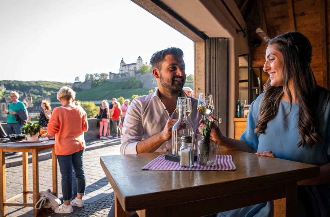 Ein junger Mann und eine junge Frau stoßen mit einem Glas Wein an. Im Hintergrund sind die Alte Mainbrücke und die Festung Marienberg im Abendlicht zu sehen.