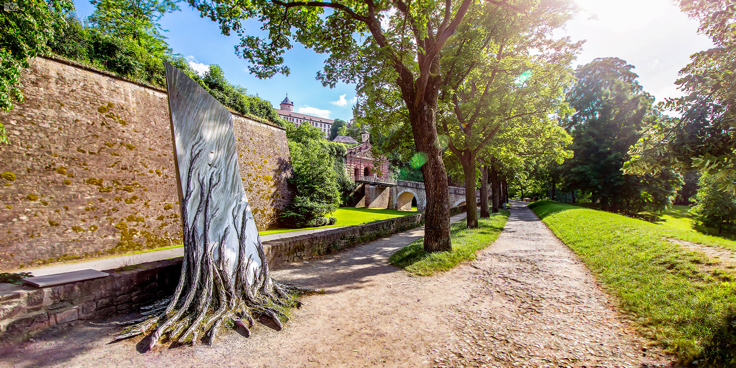 Im Vordergrund Bauerkriegsdenkmal umgeben von zahlreichen Bäumen. Im Hintergrund Festung Marienberg