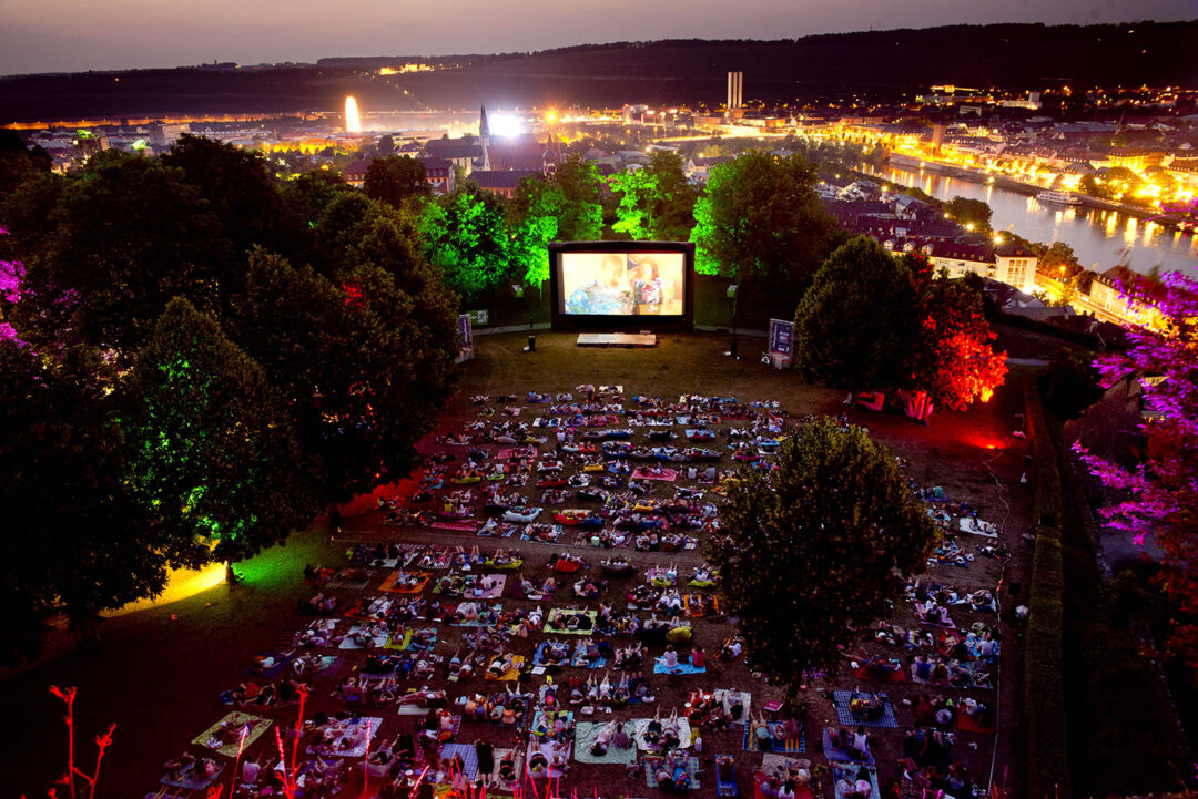 Blick von oben auf die mobile Kinoleinwand des Filmfestivals. Davor viele Besucher, die auf Decken auf der Neutorwiese sitzen.