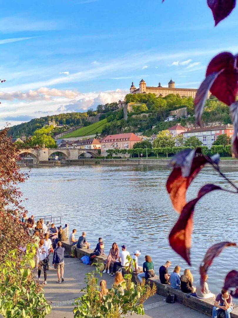 Im Hintergrund ist die Festung Marienberg zu sehen, die von der untergehenden Sonne angeleuchtet wird. Im Vordergrund sitzen viele junge Menschen auf der Kaimauer am Main